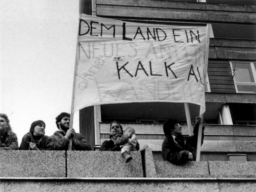 Demonstranten, die auf einem Balkon stehen. Darunter ein Kind auf dem Arm eines Erwachsenen. Zwei Demonstranten halten ein Transparent hoch. Es sind nur die Worte "Dem Land ein neues...ohne Kalk" zu erkennen.