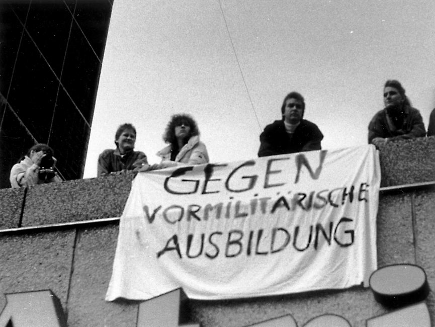 Auf dem Bild sind fünf Personen zu sehen, die auf einem Dach oder Balkon stehen. Ihre Blicke sind nach unten gerichtet. Ein Transparent mit der Aufschrift "Gegen vormilitärische Ausbildung" ist an der Fassade angebracht. Eine Person fotografiert.