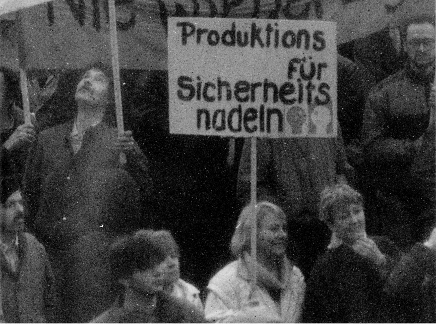 Das Bild zeigt einen Blick ganz nah in die Menschenmenge. Eine Frau hält ein Plakat mit der Aufschrift "Produktions für Sicherheitsnadeln".